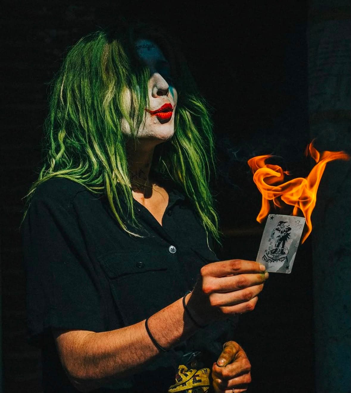 A person made up as the Joker, holding a playing card that is on fire.